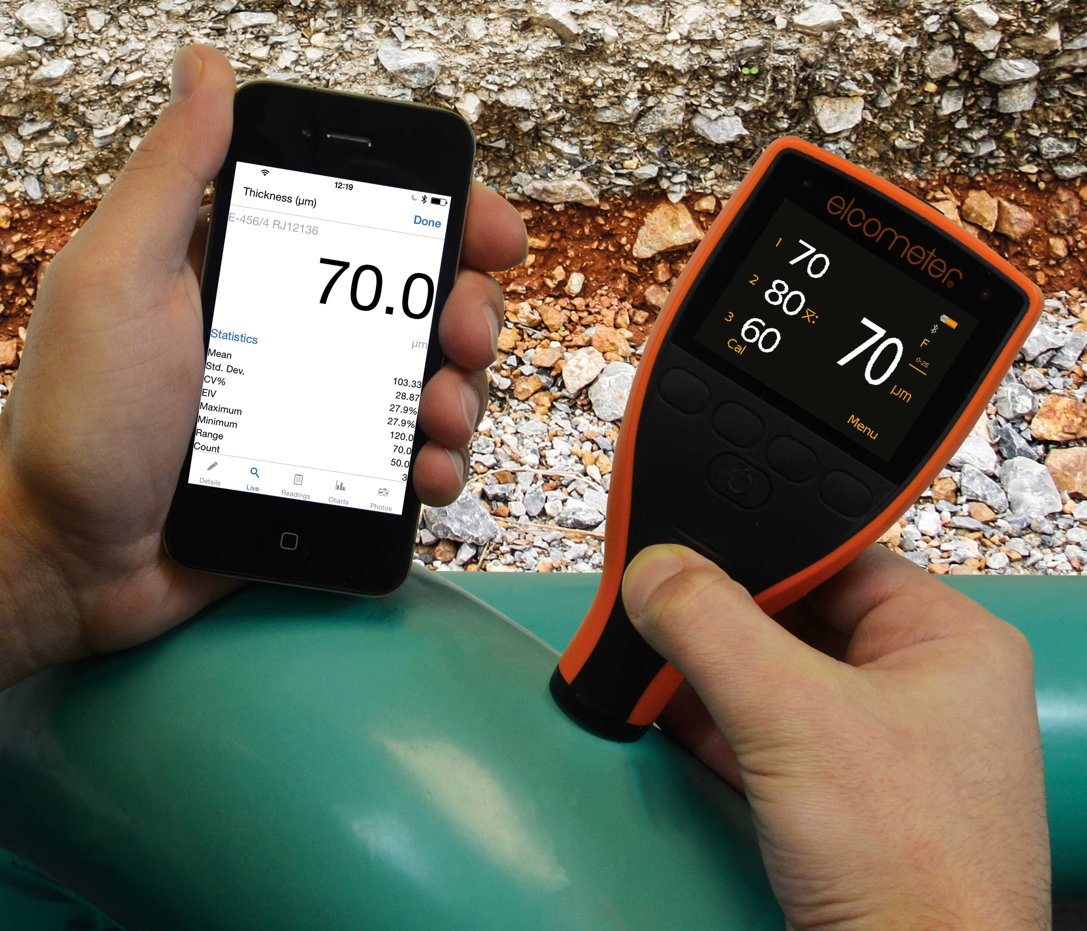 A coating inspection using the Elcometer IPC Industrial Protective Coating Thickness Gauge to measure the paint of a pipe while looking at the readings on his phone. The measurement reads '70.0'.